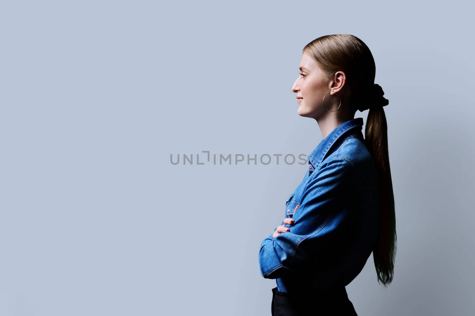 Profile view, young smiling confident female looking forward with crossed arms on gray studio background, copy space for advertising text image. Beautiful teenage blonde, fashion style beauty youth