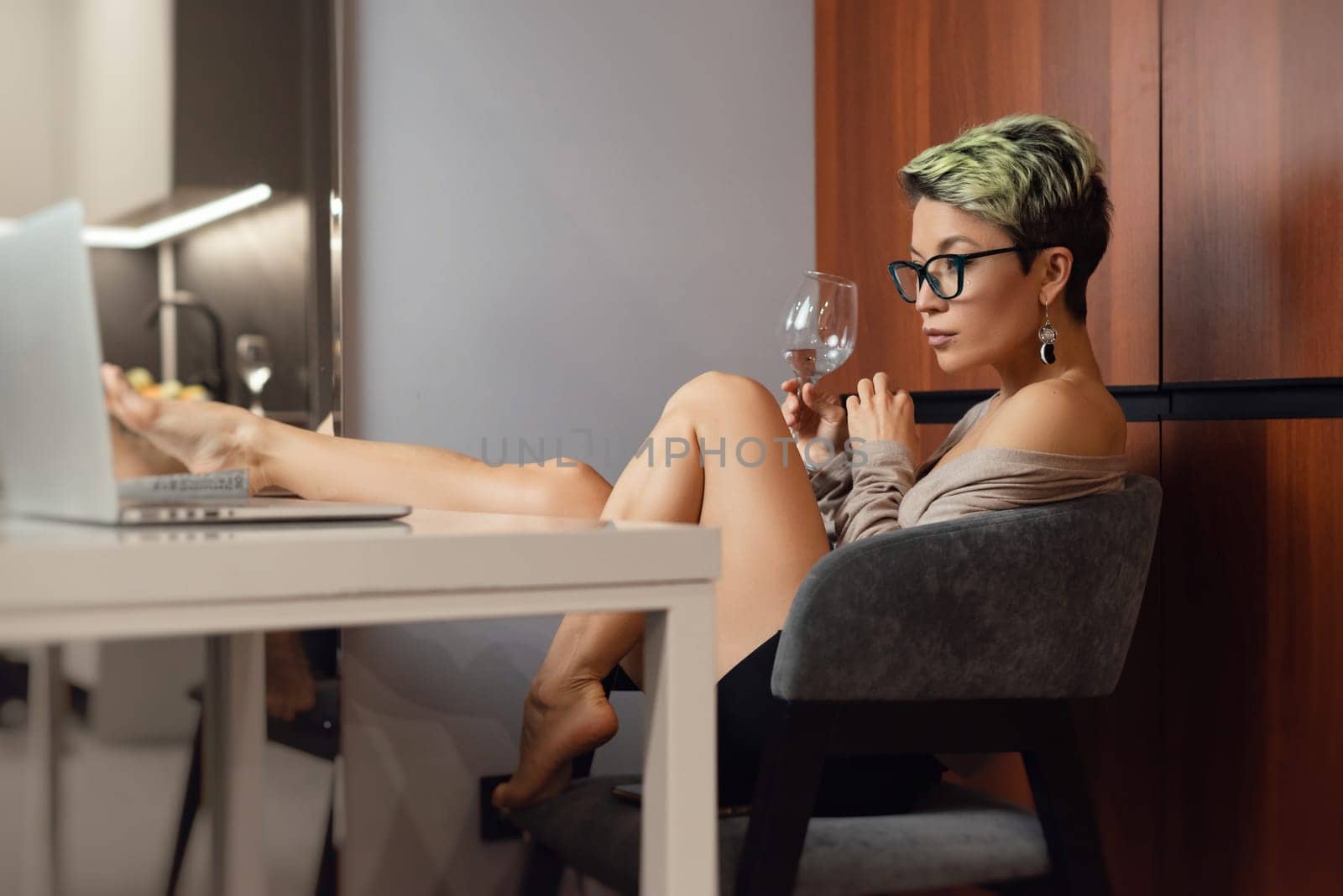 a beautiful girl with short hair and glasses is sitting indoors at a laptop, with her feet on the table, chatting and working online with a glass of water by Rotozey