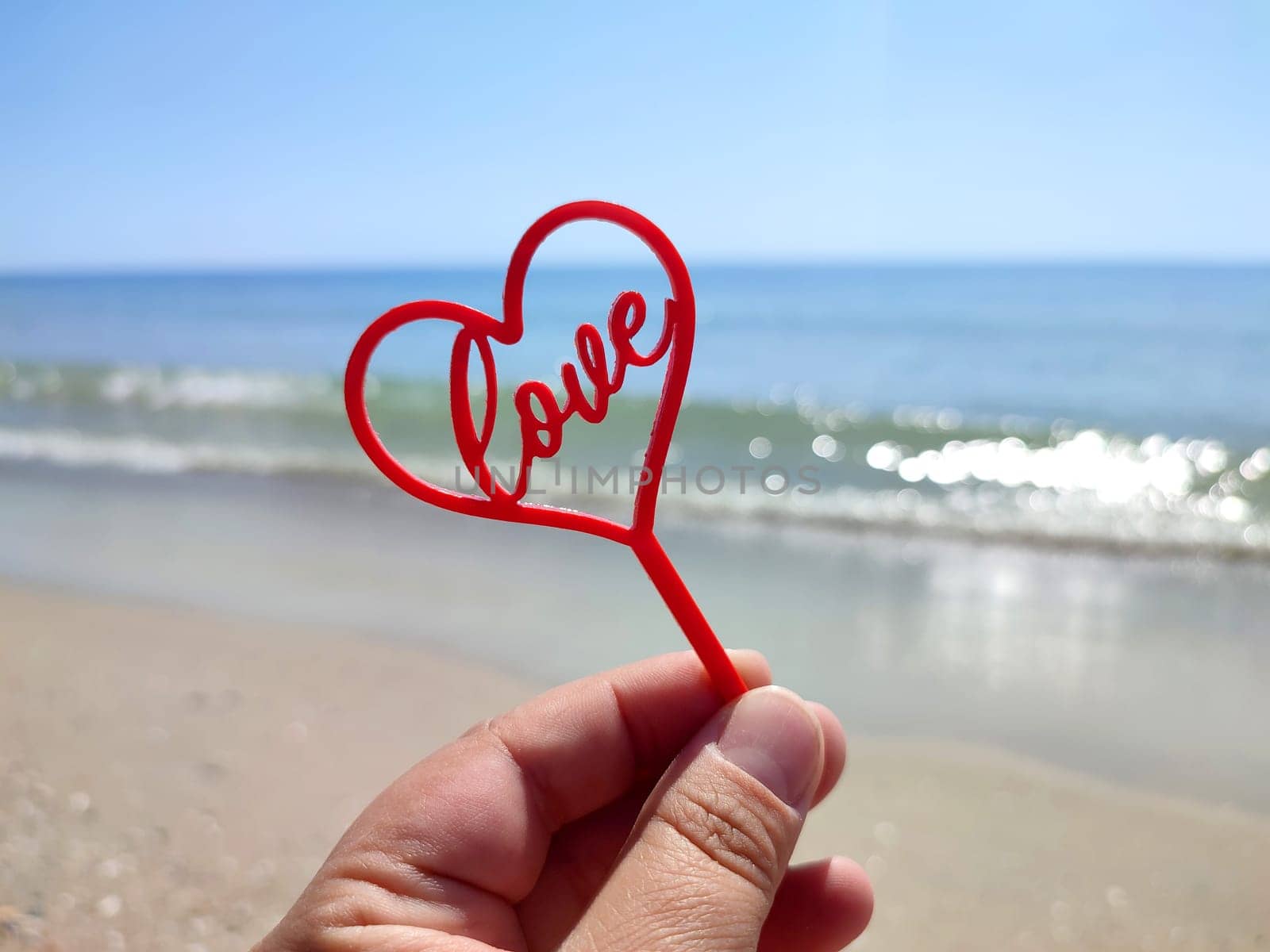 Person holding in fingers hand stick in shape red heart and word Love on background sea and sea waves, blue sky in seashore on sunny summer day close-up. Concept love romance amour St Valentines Day