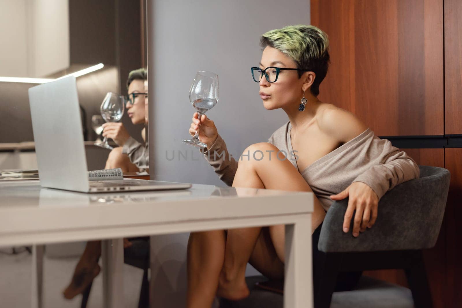 a beautiful girl with short hair and glasses is sitting indoors at a laptop, with her feet on the table, chatting and working online with a glass of water by Rotozey