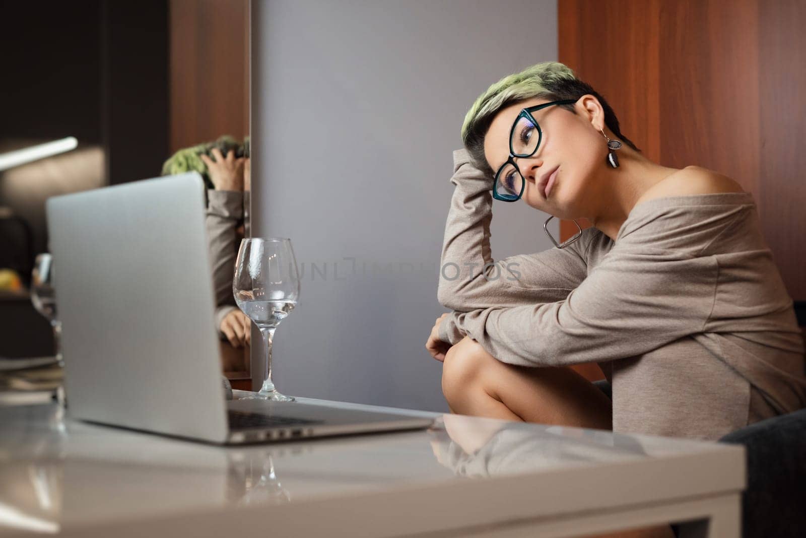 a beautiful girl with short hair and glasses is sitting indoors at a laptop chatting and working online by Rotozey