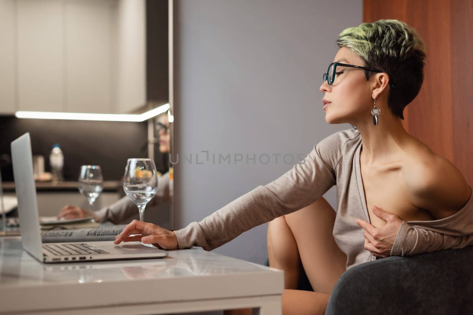 A sexy girl with short hair and glasses is sitting indoors at a laptop, chatting and working online by Rotozey