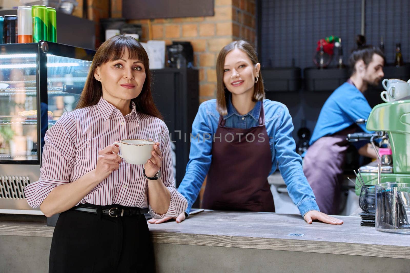 Woman customer of coffee shop near counter with cup of coffee looking at camera, restaurant workers owners at workplace. Colleagues partners in food service work, entrepreneurship, small business