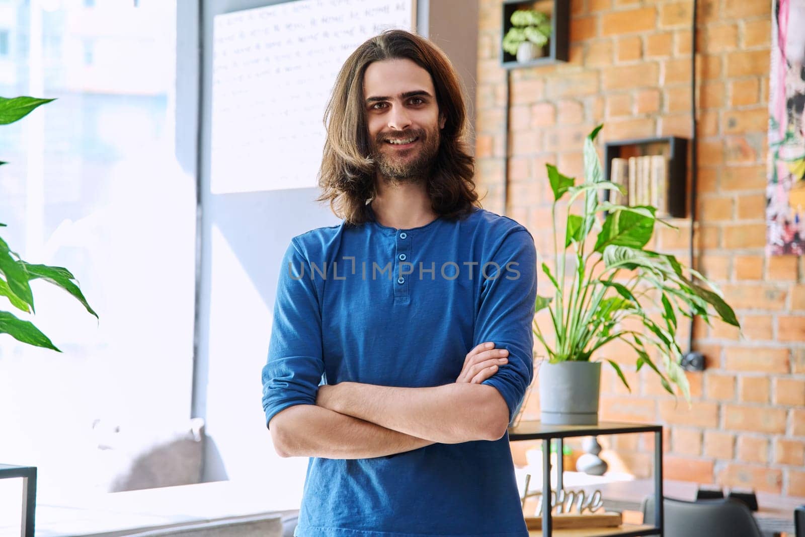 Portrait of young happy confident handsome man with long hair and beard, with crossed arms, looking at camera