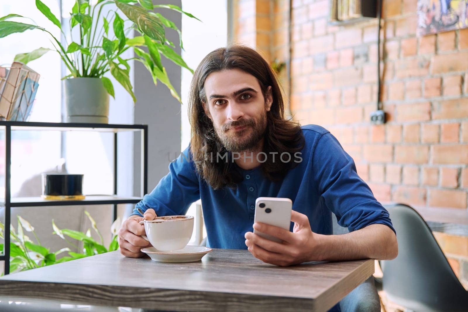 Young handsome man using smartphone, drinking cup of coffee, sitting in cafe by VH-studio