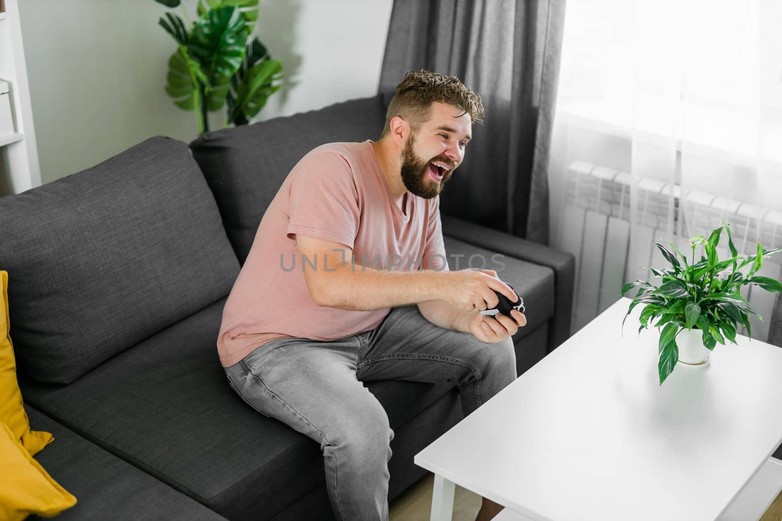 Cheerful young man seated on a sofa playing video games at home. Gaming addiction and entertainment by Satura86