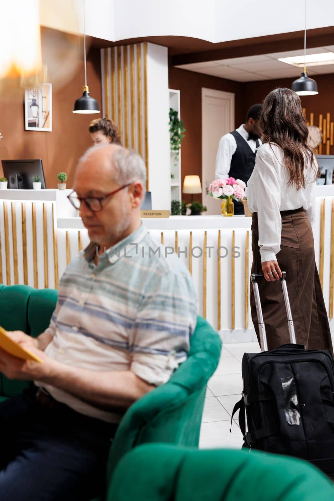 Caucasian elderly lady with luggage enters contemporary hotel lobby and proceeds to front desk to check in. Senior female tourist with suitcase approaches counter for accommodation booking procedure.