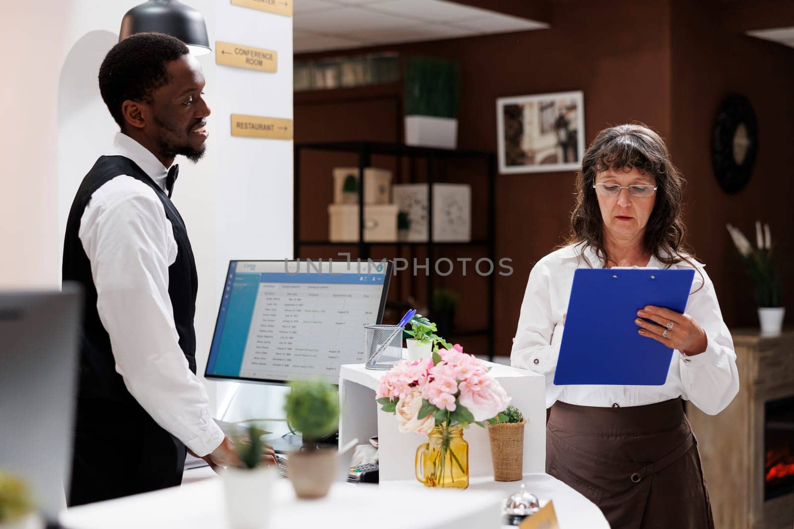 Elderly female tourist checks in at hotel reception, ready for enjoyable holiday. Retired senior woman fills out room reservation paperwork with help from african american male receptionist.