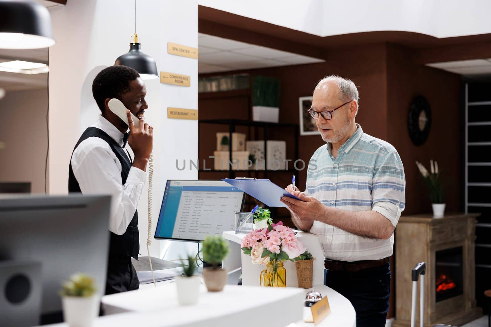 Caucasian retired old man signs reservation documents at hotel front desk as african american concierge answers call. Eldery male client filling check-in forms while receptionist speaks on telephone.