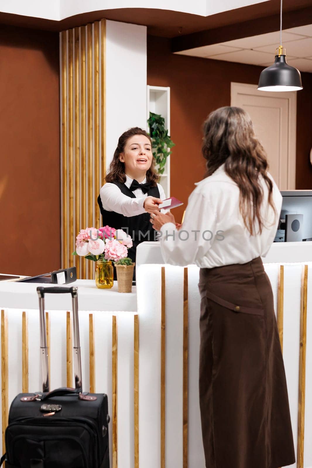 Elderly female tourist checks in with her passport at hotel lobby desk. After confirming identity, friendly concierge at registration counter returns the personal documents of retired senior woman.