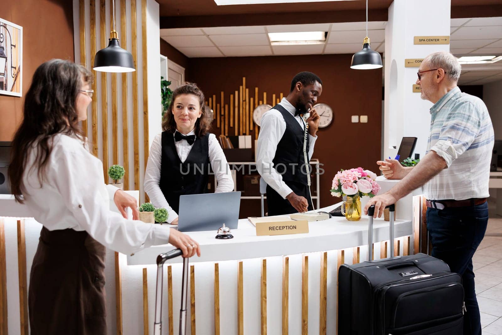 Two elderly travelers come at the hotel reception desk to check in. Multiethnic receptionists assist retired senior man and woman with booking and paperwork, therefore creating a pleasant environment.