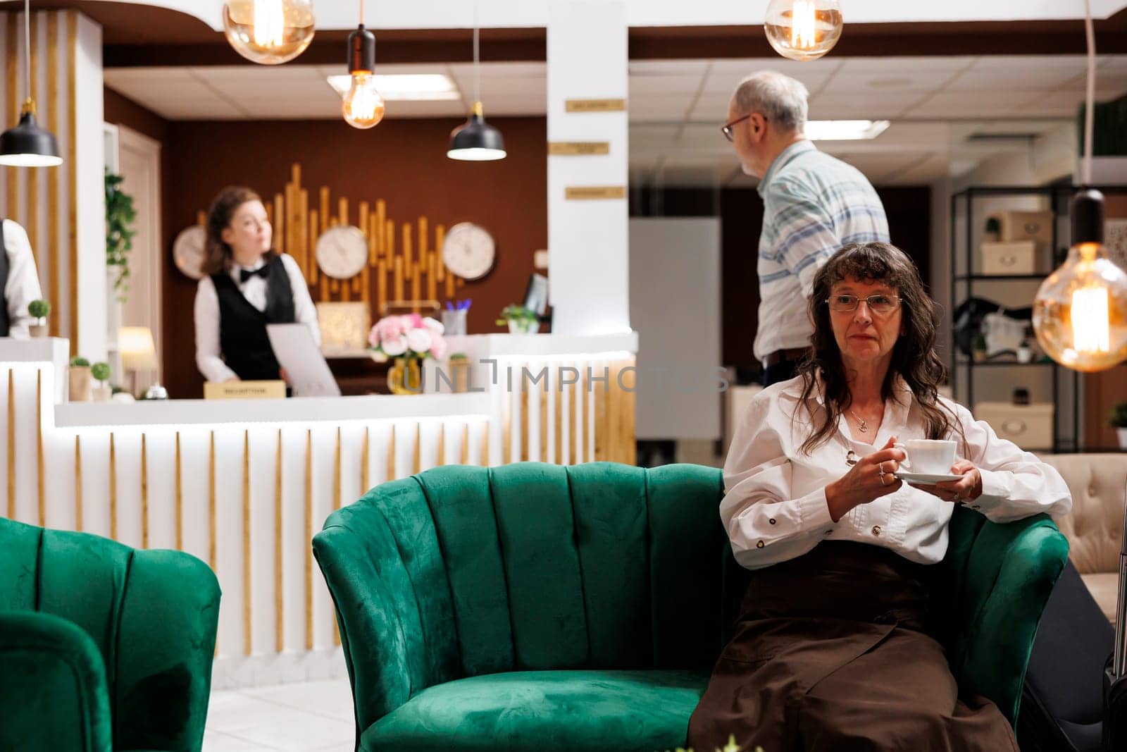 Elderly caucasian woman seated on sofa with cup of coffee at luxurious lounge area for check-in. Senior man arrives in hotel lobby while old female tourist waits on couch for booking procedure.