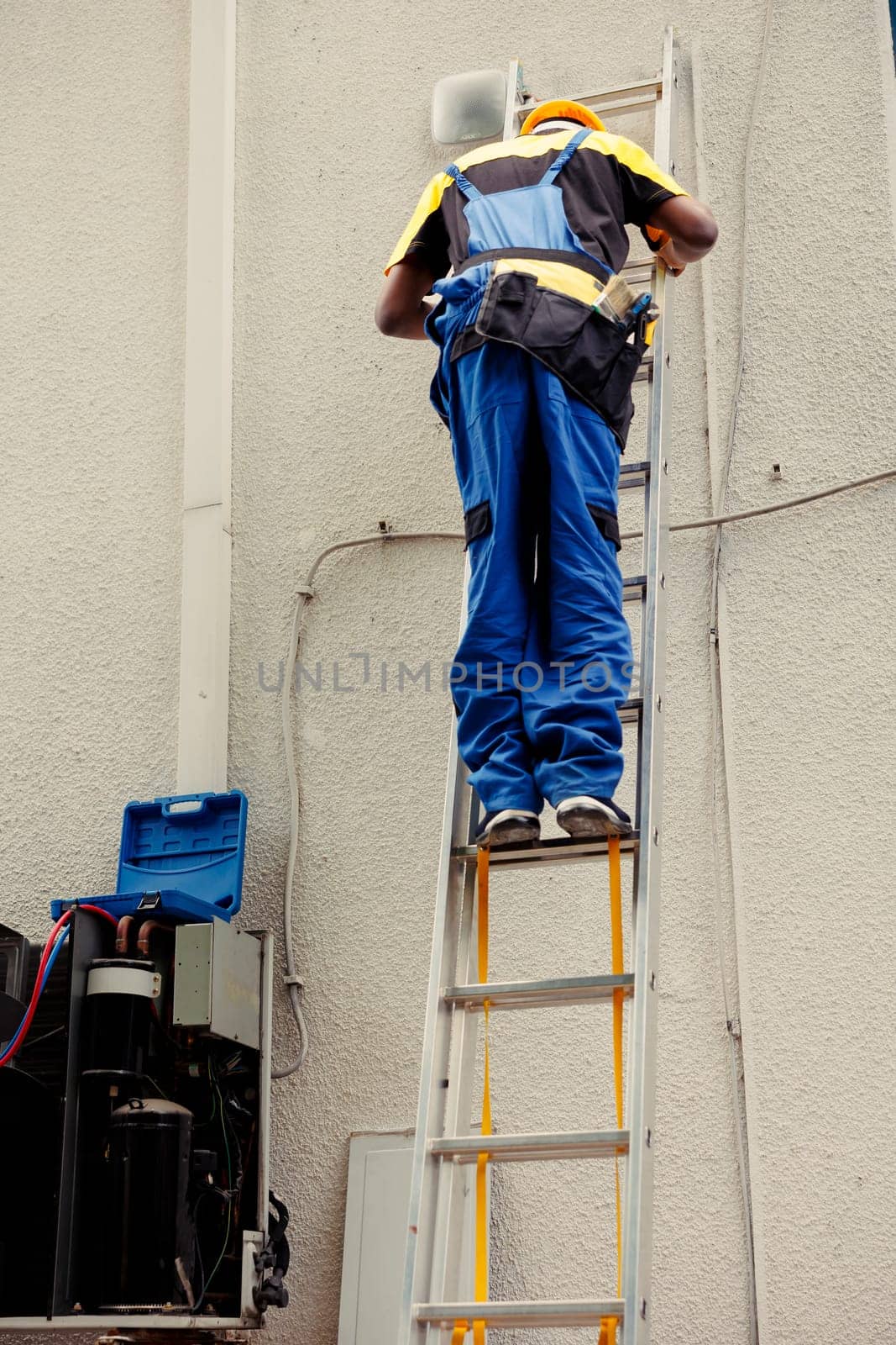 Competent mechanic in professional uniform with technical equipment climbing folding ladder to do maintenance on rooftop hvac system. Expert repairman commissioned to check up on air conditioner
