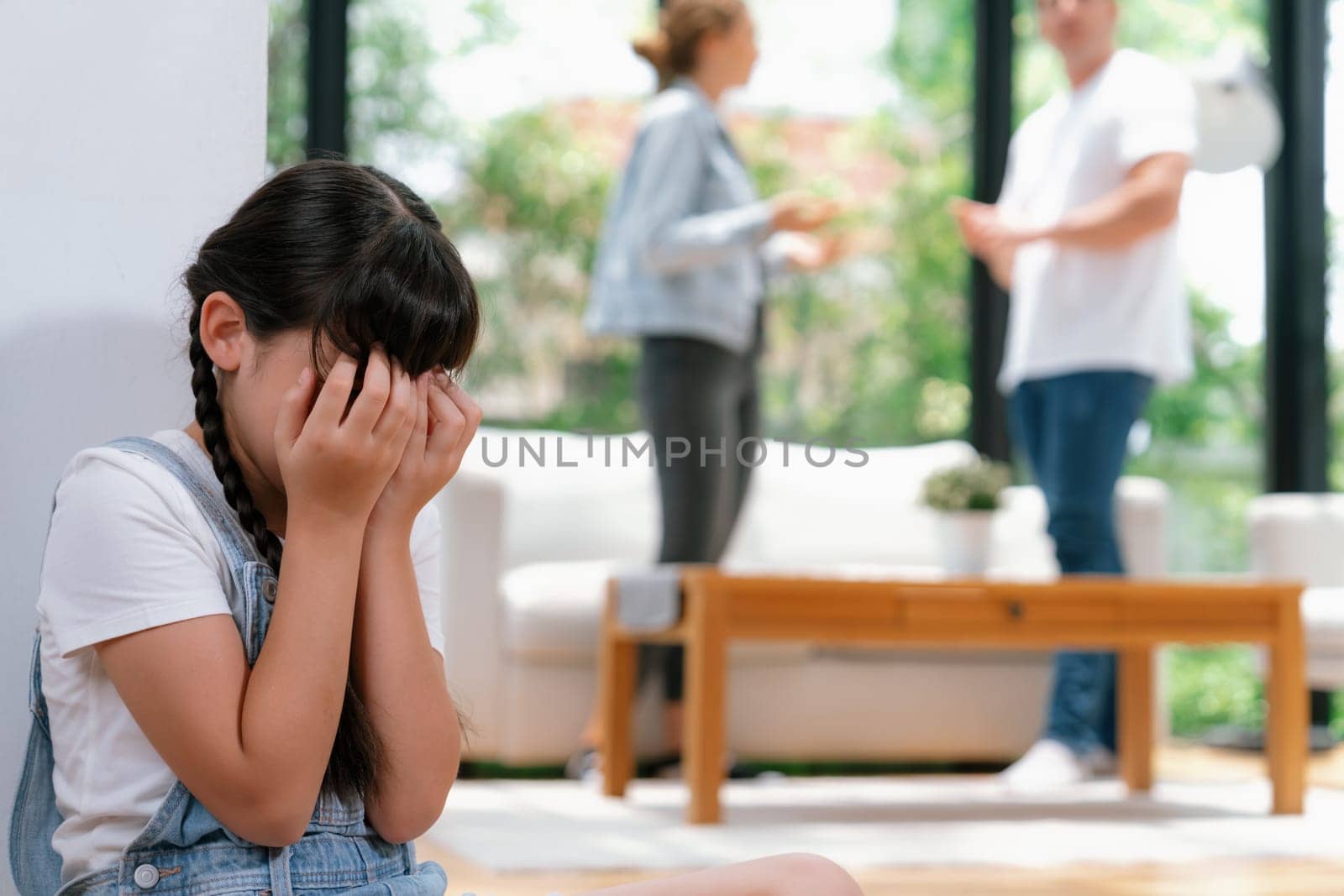Stressed and unhappy young girl huddle in corner, cover her face while her parent arguing in background. Domestic violence at home and traumatic childhood develop to depression. Synchronos