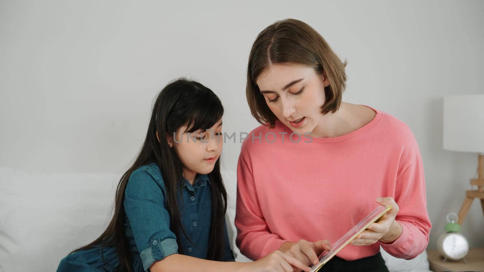 Caucasian mother reading story book to cute daughter listening story. Asian girl pointing while talking about story before bed time. Happy family sitting on bed while spend time together. Pedagogy.