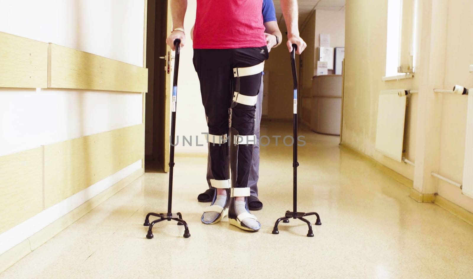 Legs of disable man in orthosis walking with support of two walking cane in the rehabilitation clinic. Doctor helping him.