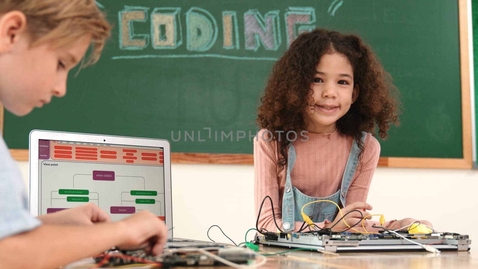 Girl standing while fixing electronic board by using screwdriver. American student and happy caucasian boy working together to inspect electric system. Curious children working on board. Pedagogy.