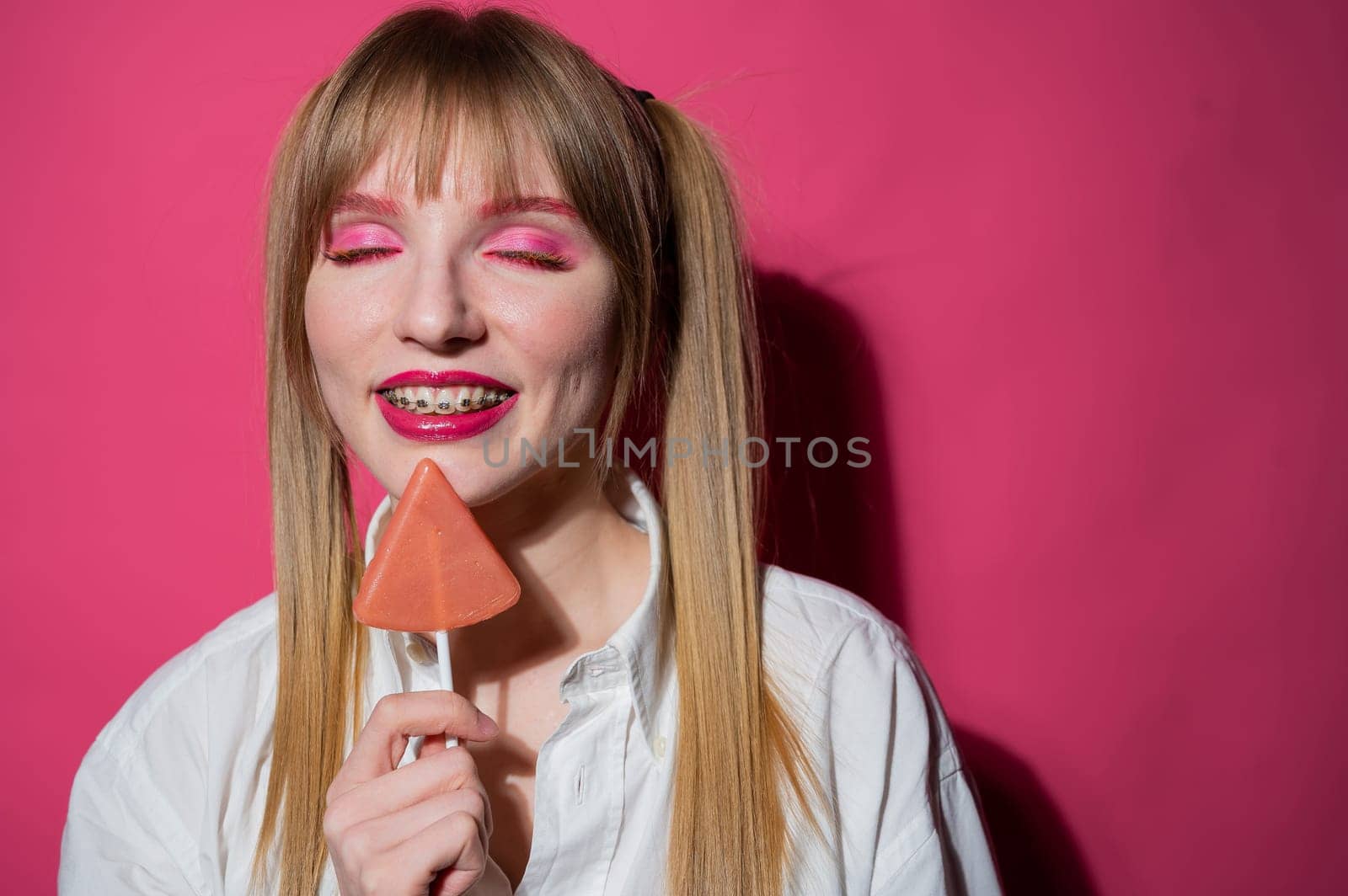 Portrait of a young woman with braces and bright makeup eating a lollipop on a pink background. by mrwed54