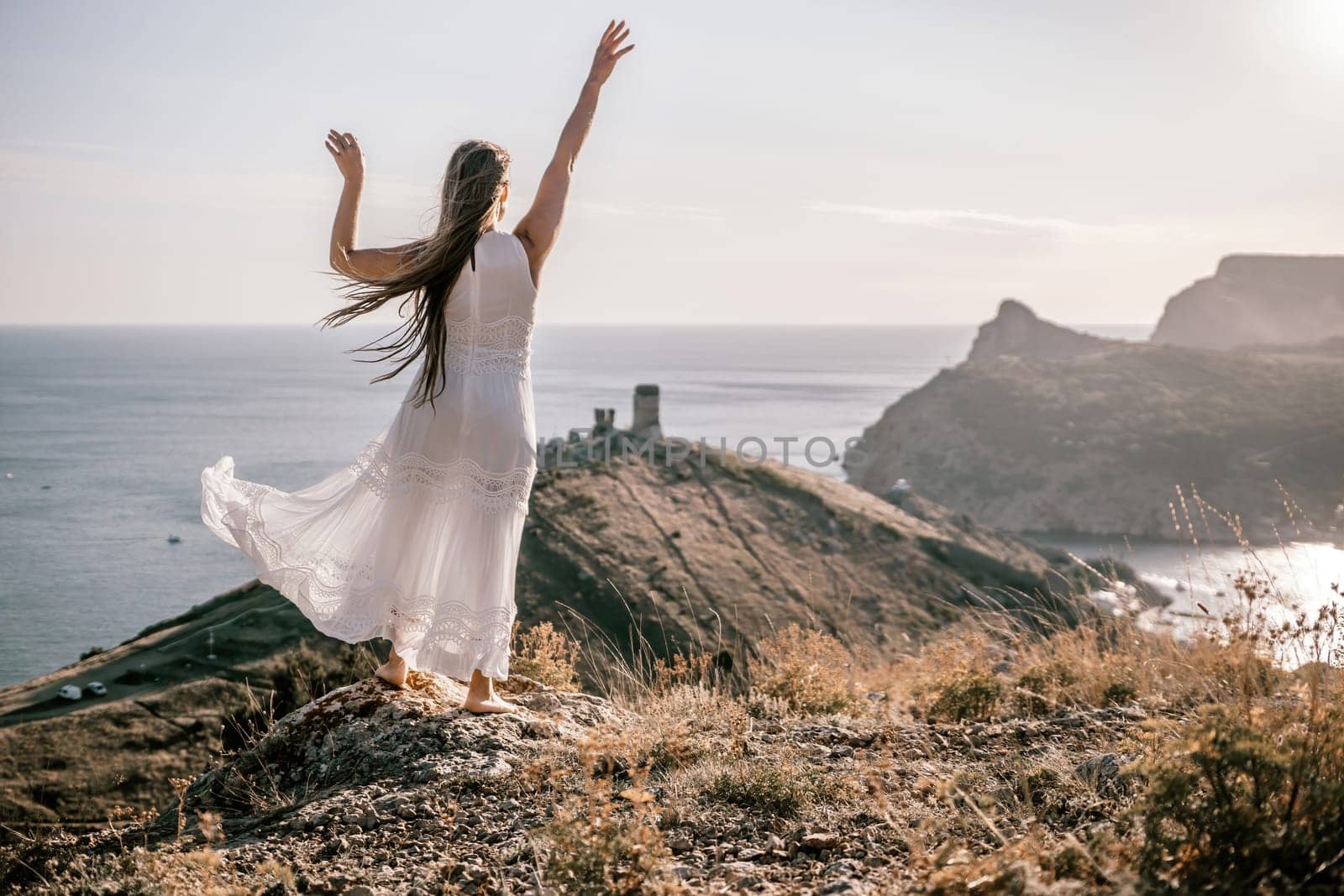 A woman in a white dress stands on a rocky hill overlooking the ocean. She is smiling and she is happy. by Matiunina