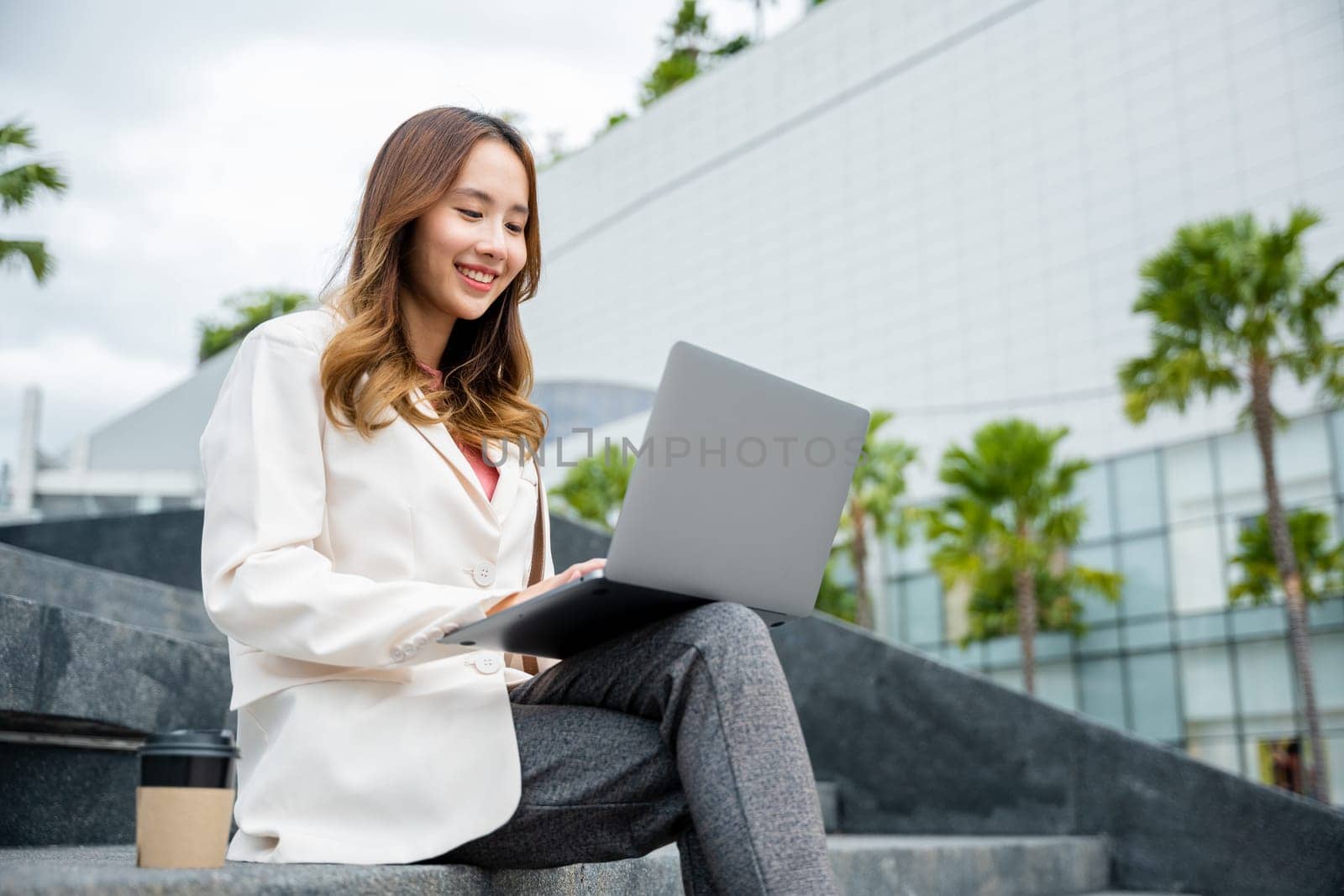 woman freelance smile using laptop computer with smile while working with coffee cup while sitting on stairs outdoors by Sorapop
