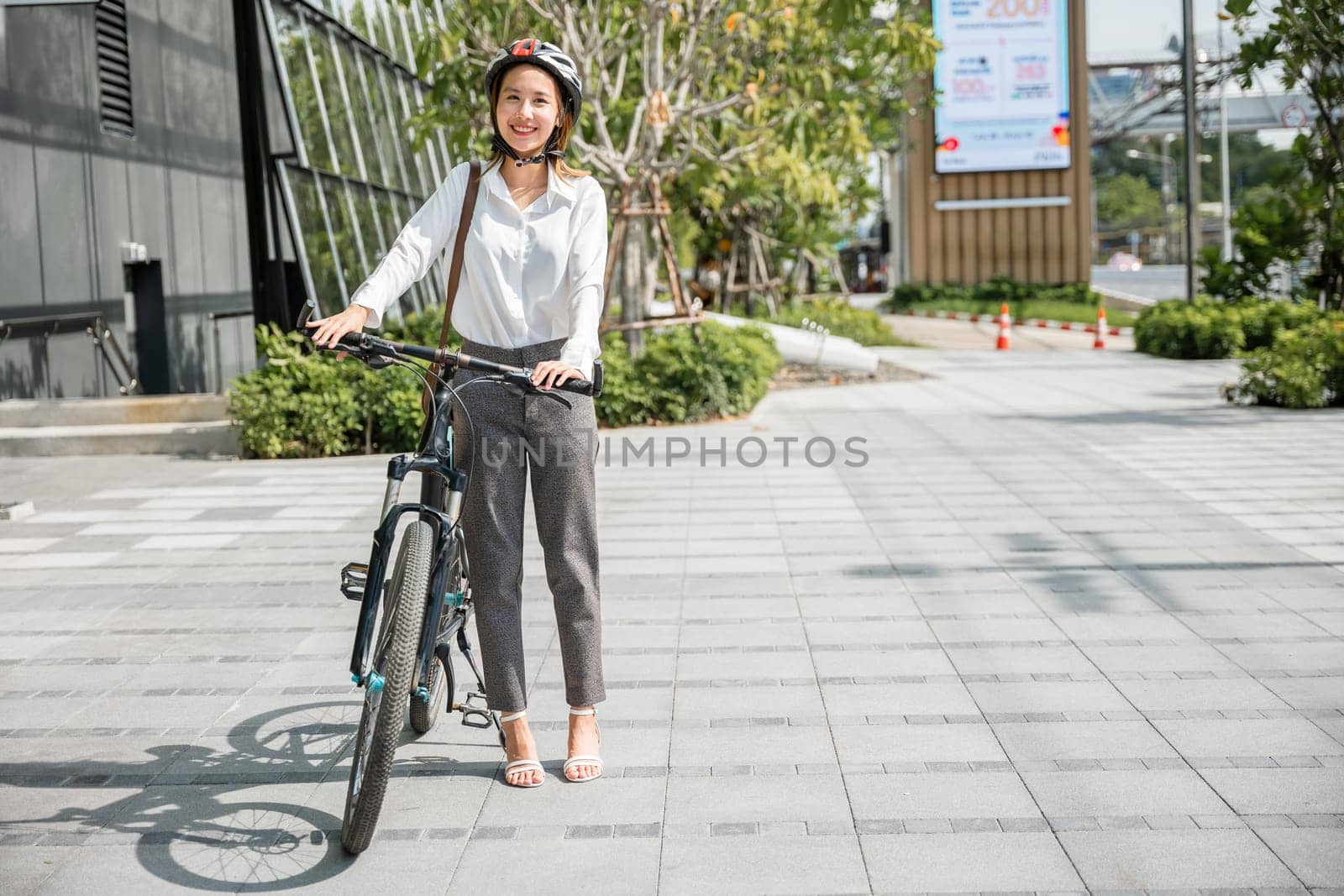 In a city setting Asian businesswoman in a suit and helmet smiles beside her bicycle ready for her morning commute. portrays the cheerful blend of work and outdoor activity in business commuter life. by Sorapop