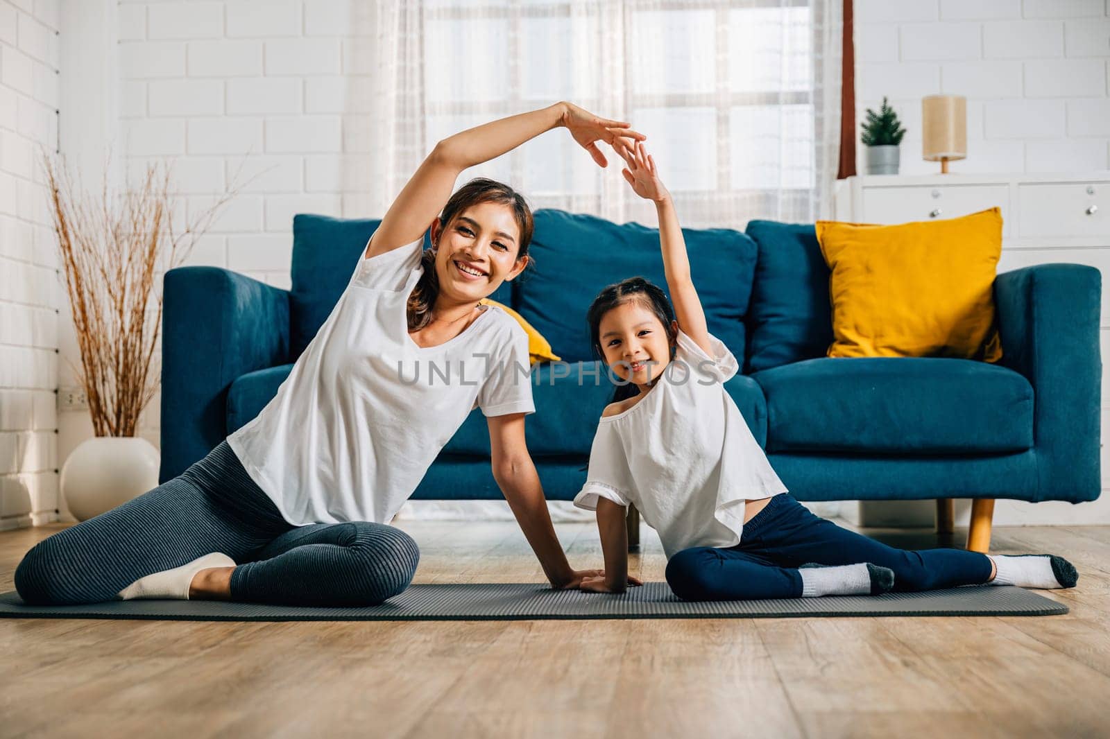 In their home an Asian mother becomes a yoga teacher for her daughter promoting strength harmony and education. The smiles and concentration are evident in this family's happy moment.