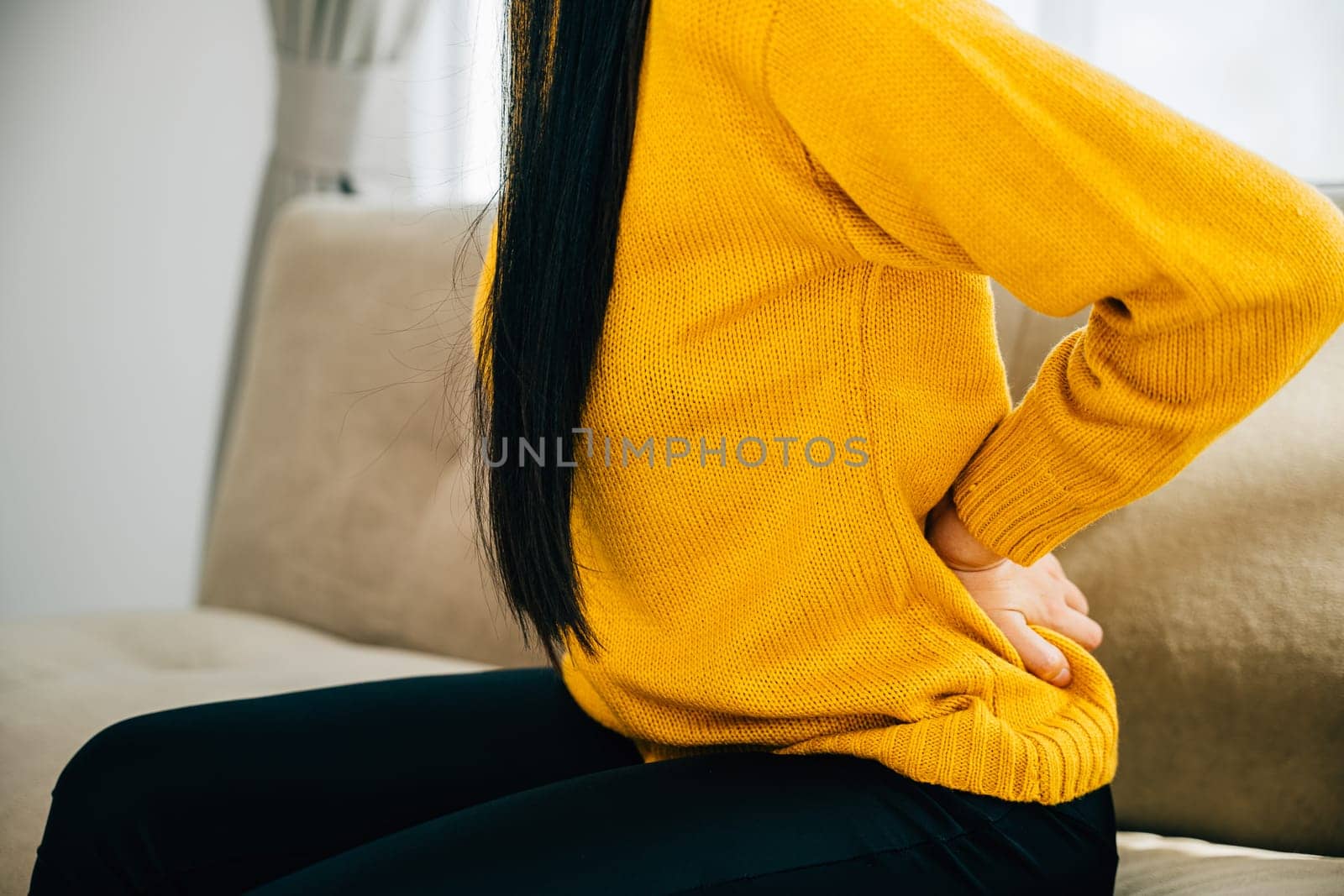 A woman from Asia holds her lower back in unbearable pain while seated on a sofa. Emphasizing chronic back pain discomfort and importance of medical care for back issues. health problem concept