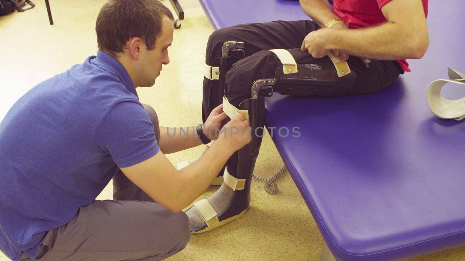 Doctor physiotherapist putting the orthosis on the legs of young disable man in the rehabilitation clinic. Patient helping him