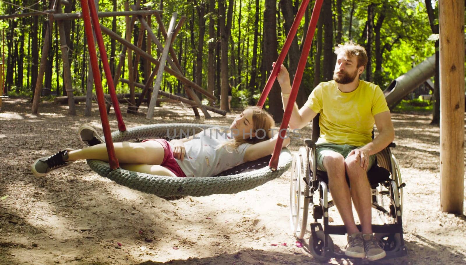 Happy young disable man in a wheelchair in the park with his wife. Woman is lying on a swing and a man is pushing a swing