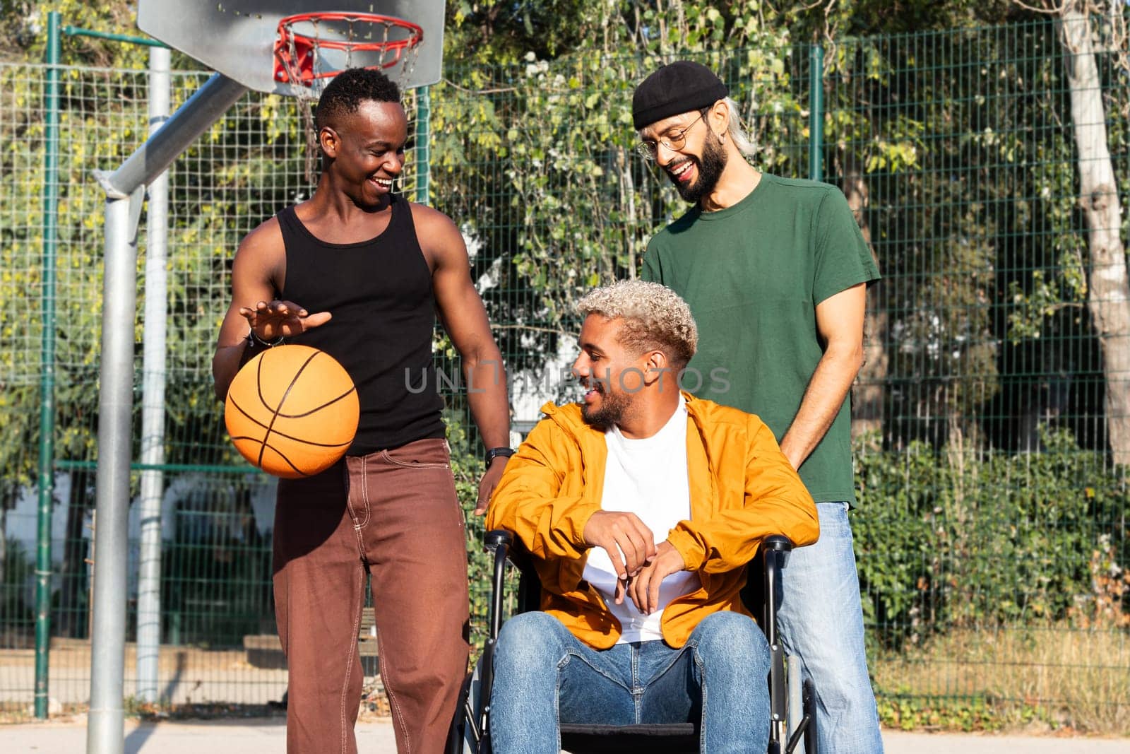 Young Black man in a wheelchair talking and laughing with friends in basketball court outdoors. Sports and friendship concept.