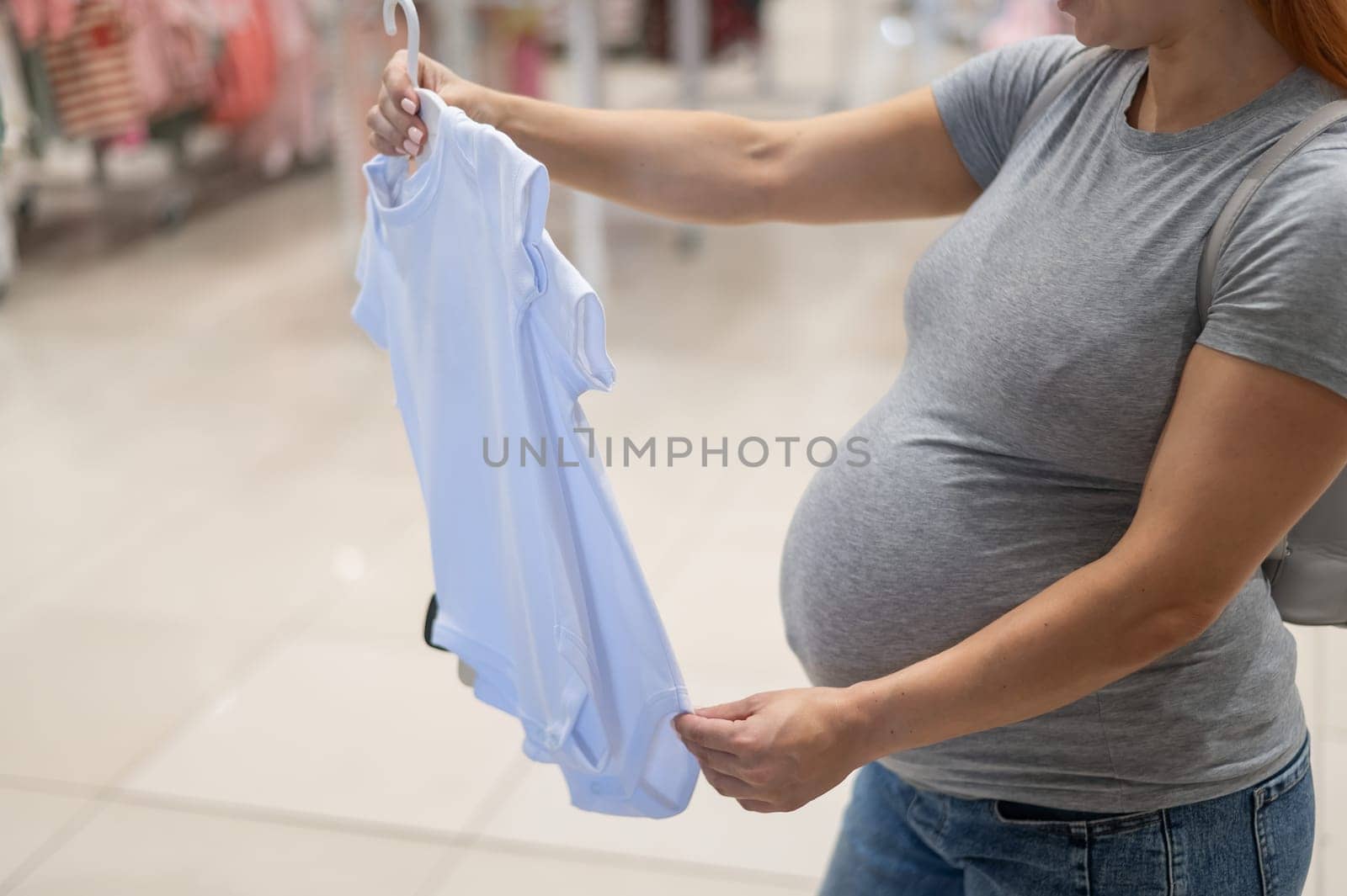 Caucasian pregnant woman chooses baby clothes in a store. Faceless expectant mother in the 3rd trimester