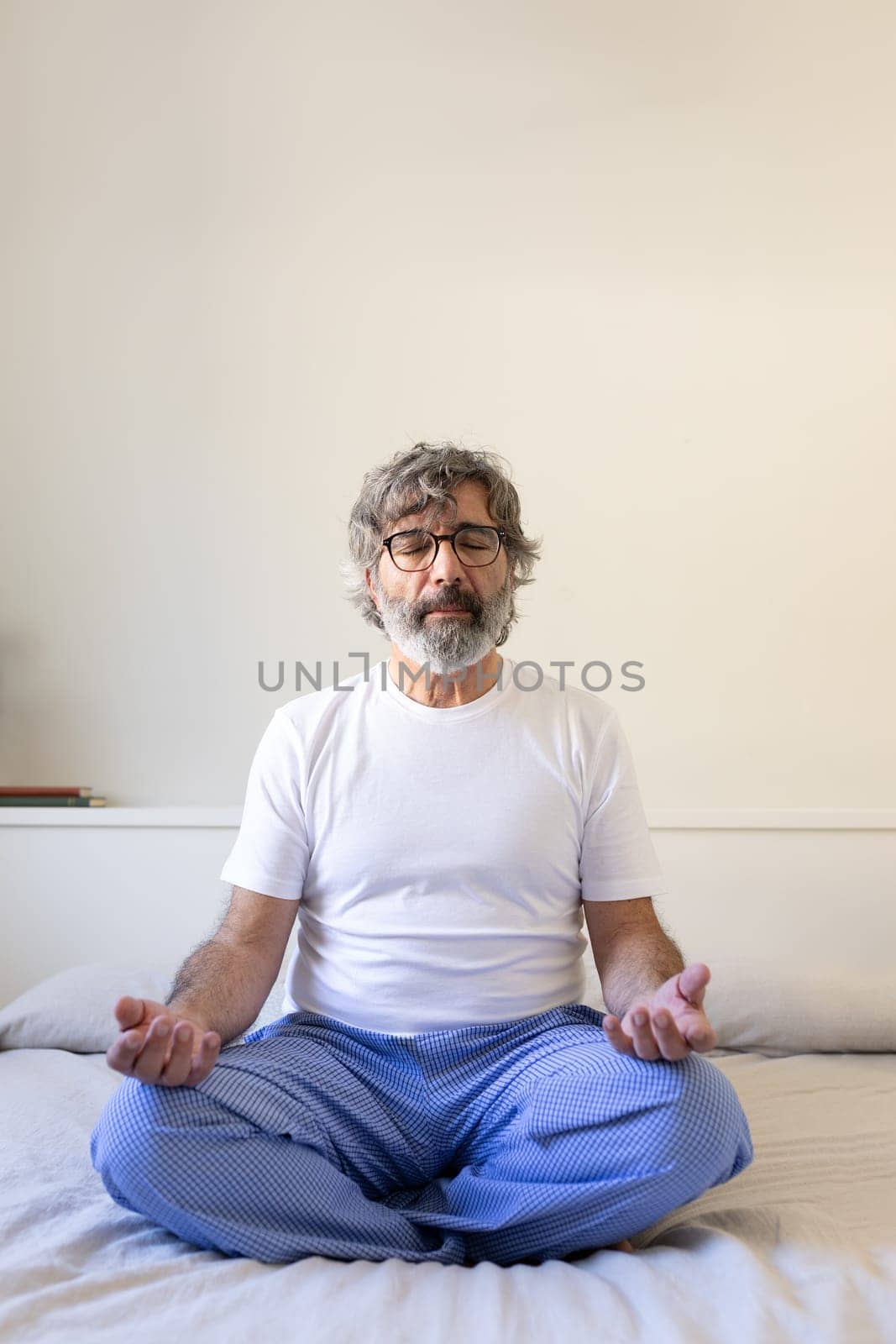 Front view vertical portrait of mature adult man relaxing, doing meditation sitting on bed at home. Male meditating in the morning. Wellness and mental health concept.