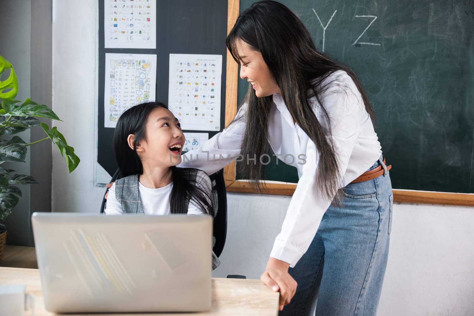 A woman is smiling at a young girl. The woman is the teacher and the girl is her student. The scene is set in a classroom with a blackboard and a laptop computer