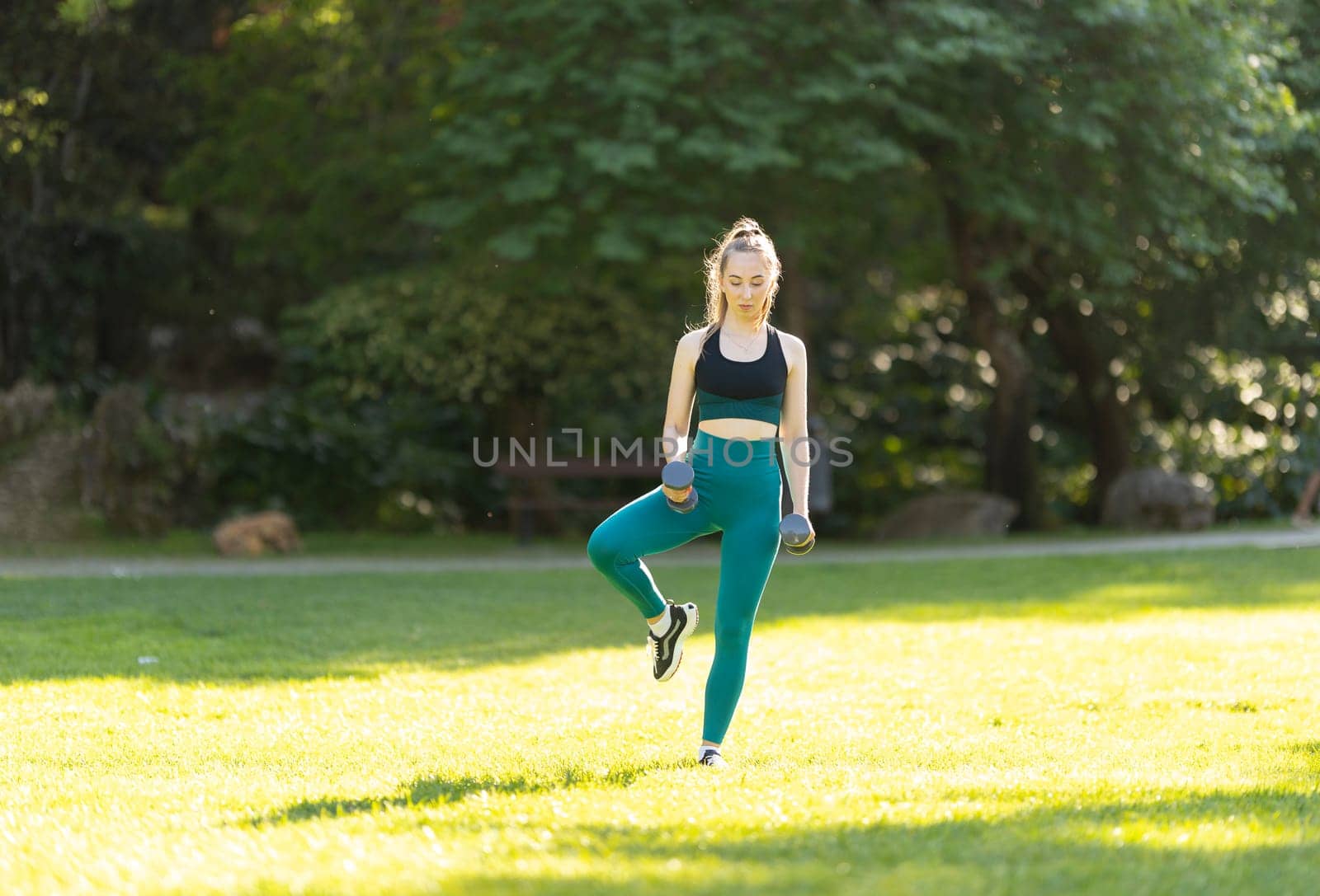 A woman is exercising in a park. She is wearing a green top and blue pants. She is holding two dumbbells in her hands