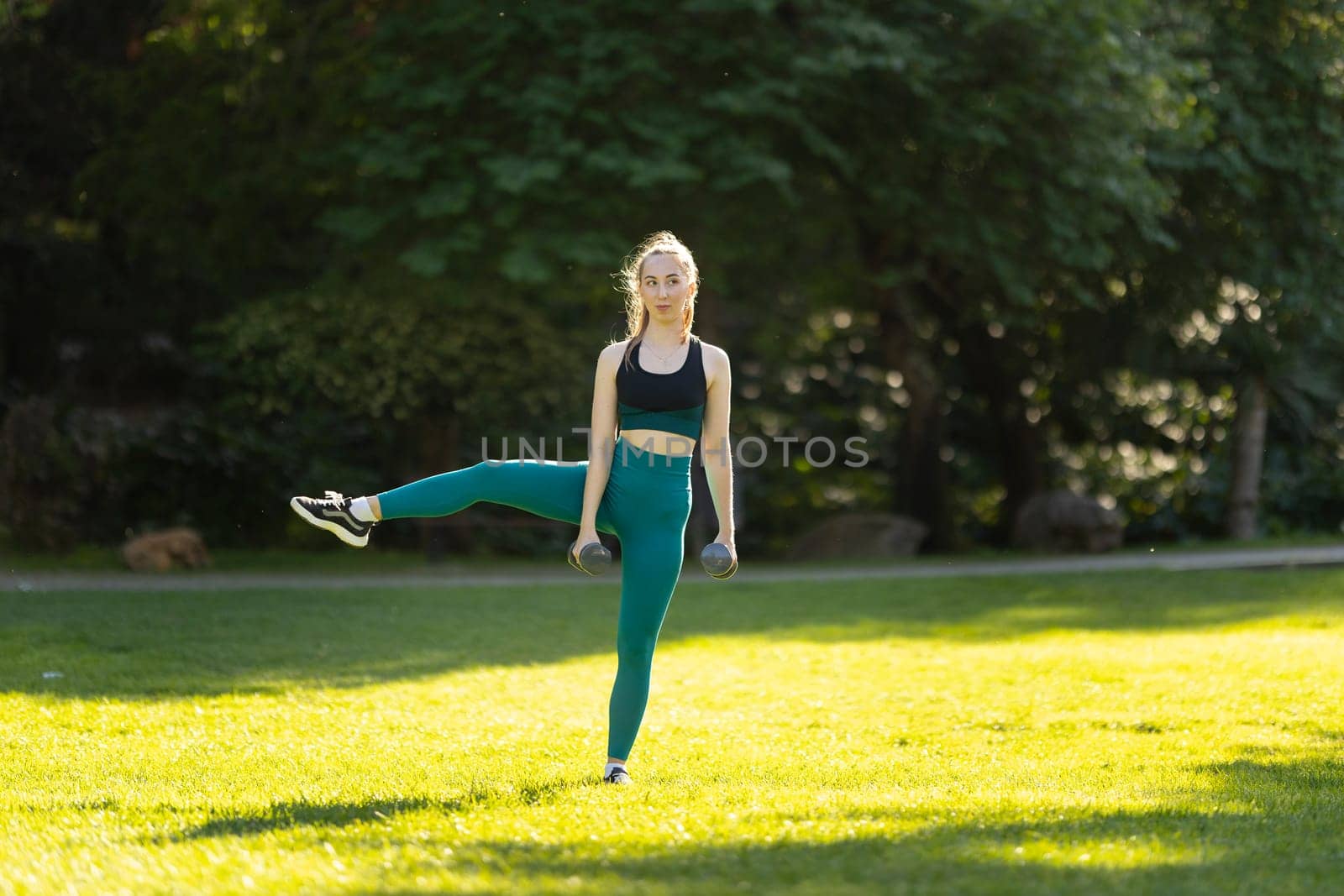 A woman is doing a leg lift in a park. She is wearing a black tank top and green leggings