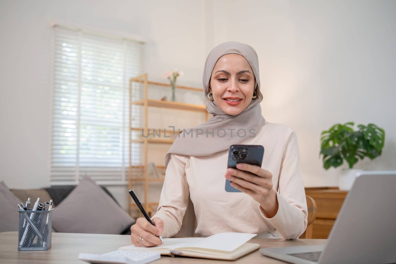 A young Muslim woman wearing a hijab calculates financial accounts. Income and expenses in the living room at home by wichayada