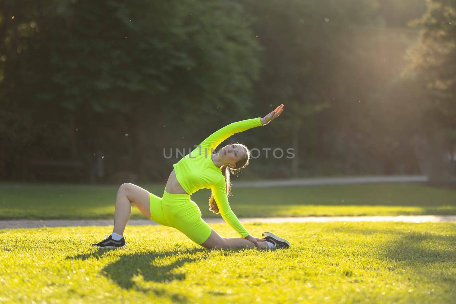 A woman in a neon green top and shorts is doing a yoga pose on a grassy field, sunset