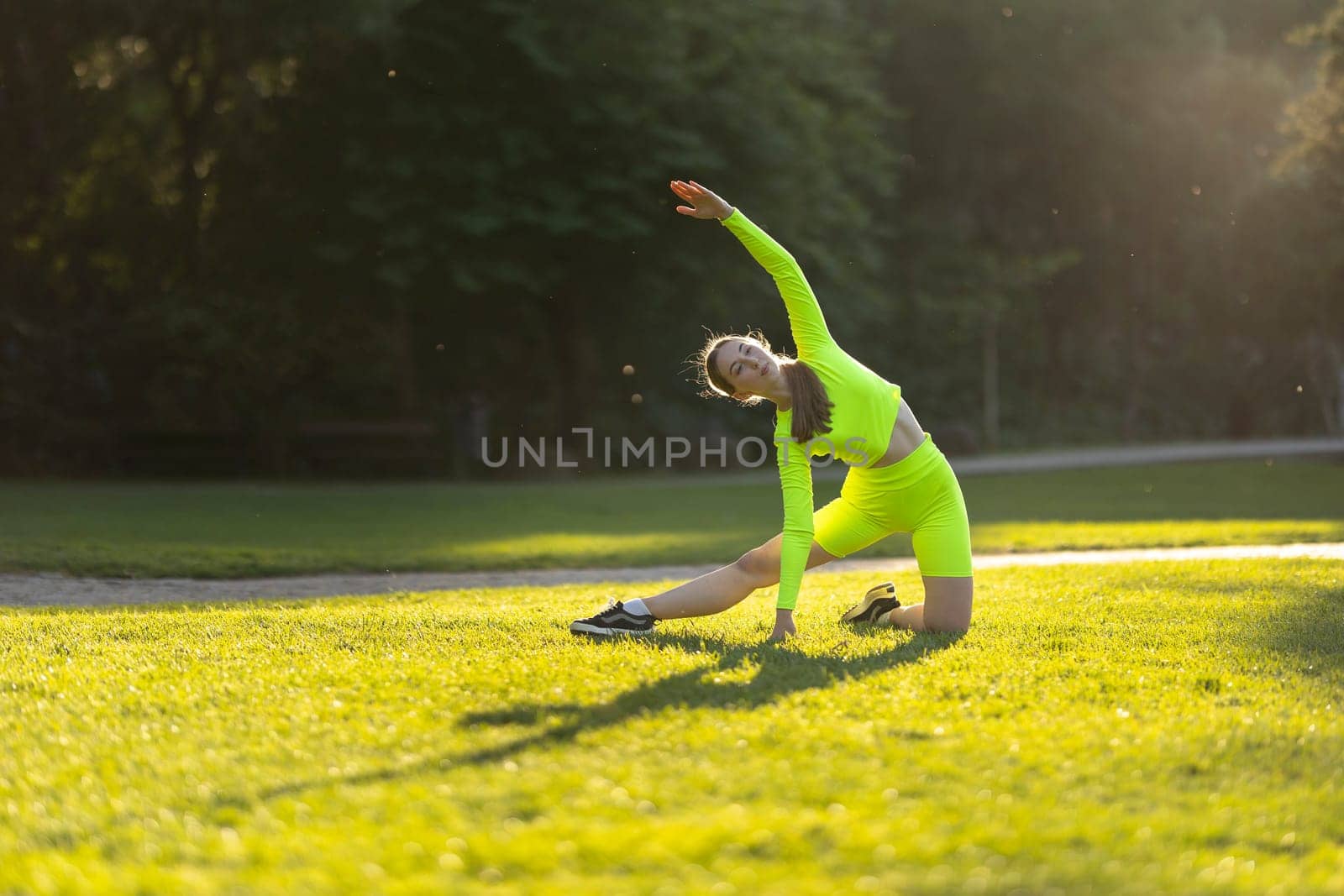 A woman in a neon green outfit is doing a yoga pose on a grassy field. The bright colors of her outfit and the surrounding green grass create a cheerful and energetic atmosphere