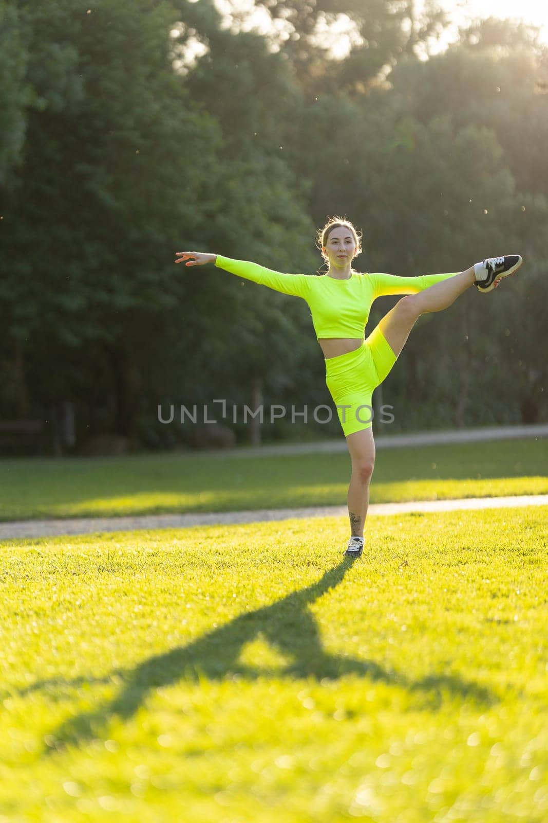 A woman in a neon yellow outfit is doing a split on a grassy field. Concept of energy and athleticism, as the woman's pose