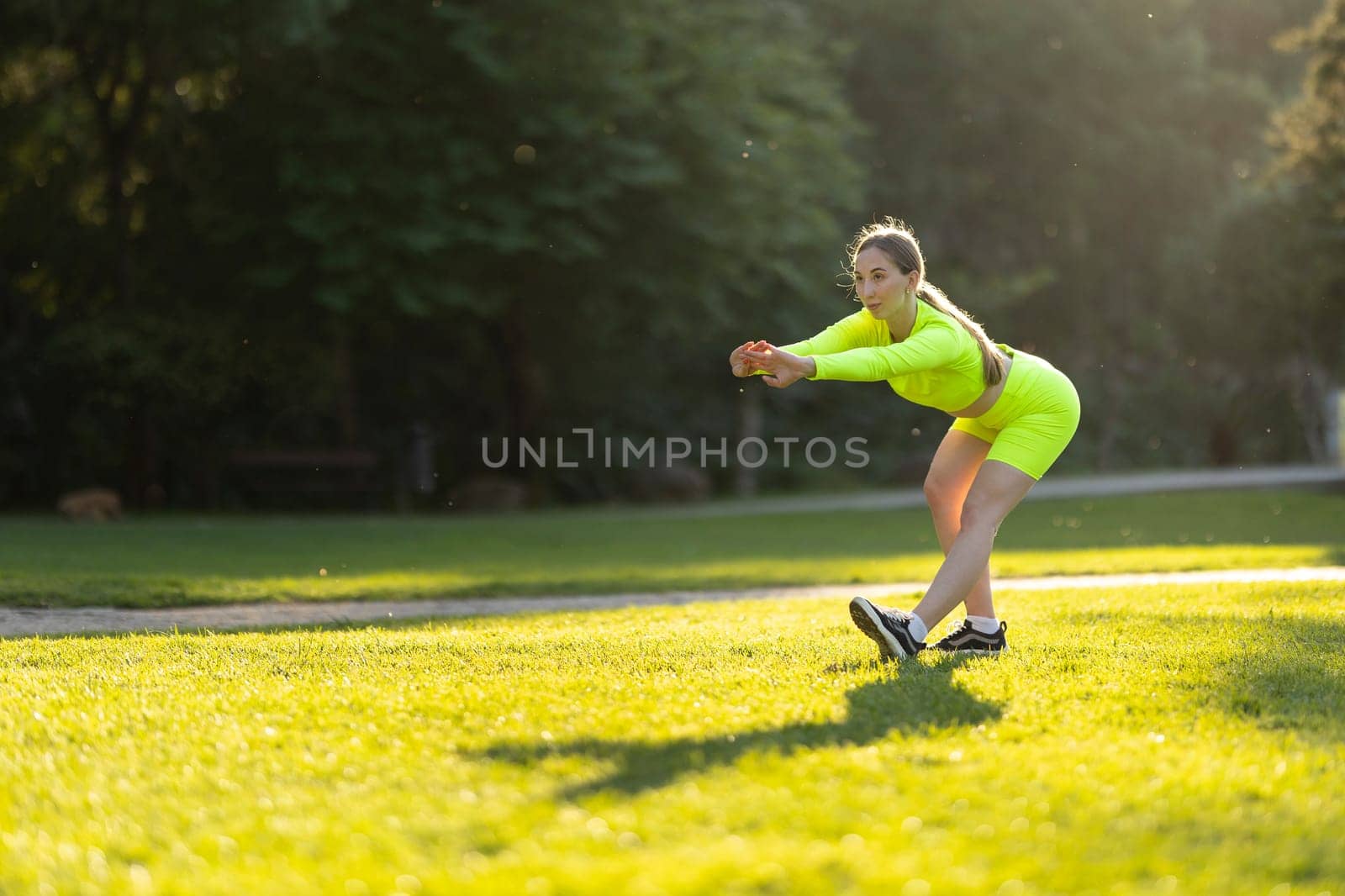 A woman in a neon yellow outfit is stretching on a grassy field by Studia72