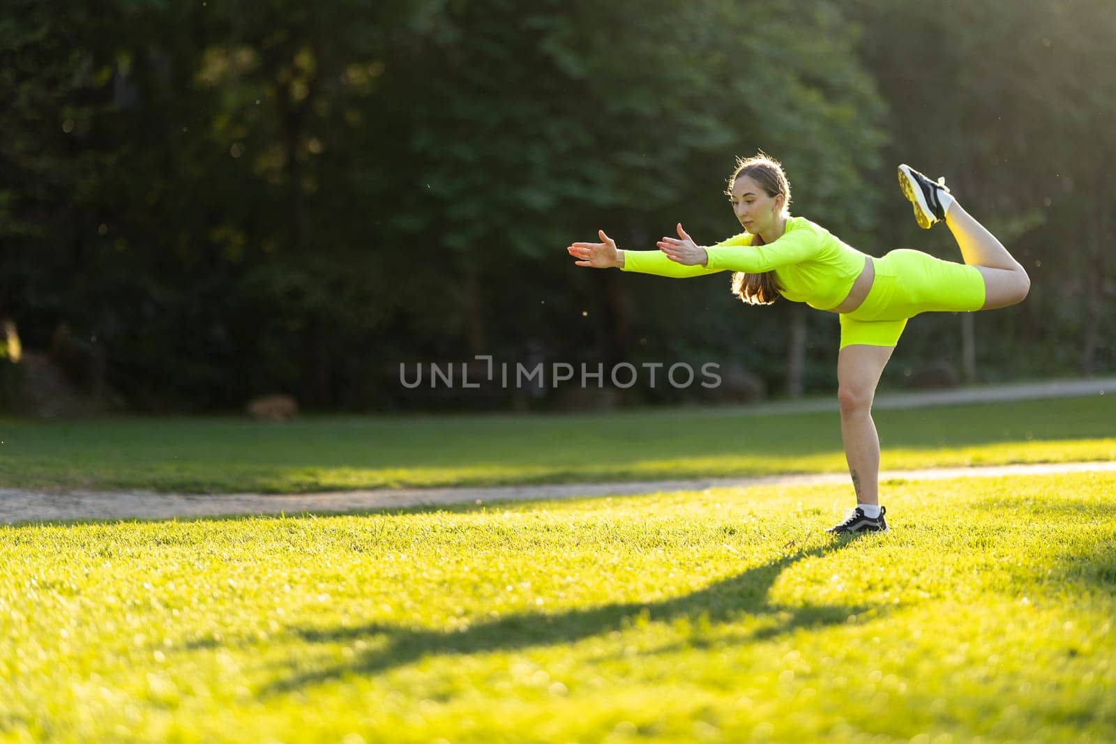 A woman is doing a yoga pose on a grassy field. The grass is green and the sky is blue