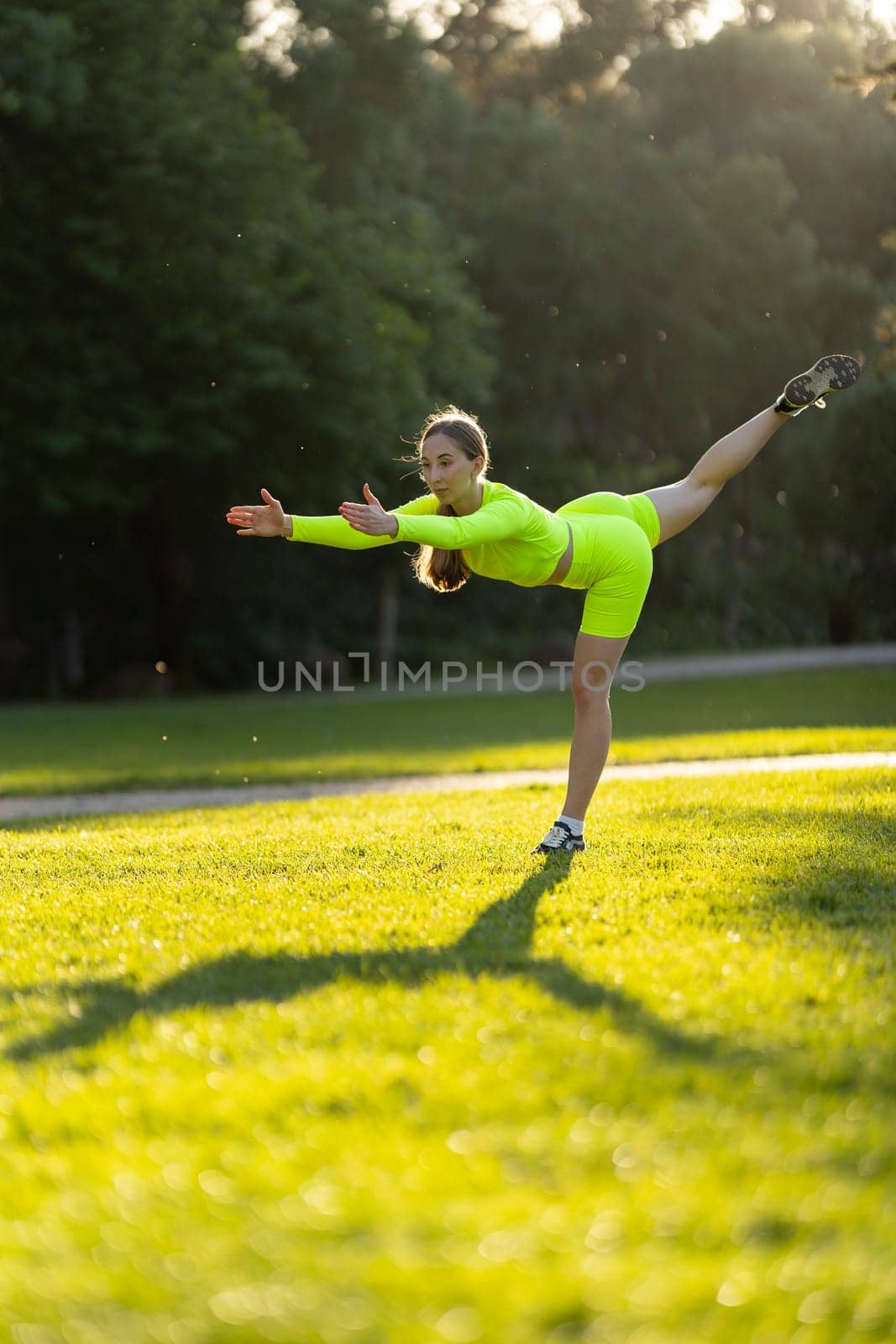 A woman in a neon yellow outfit is doing a yoga pose on a grassy field. The bright colors of her outfit and the green grass create a cheerful and energetic mood. Concept of health and fitness
