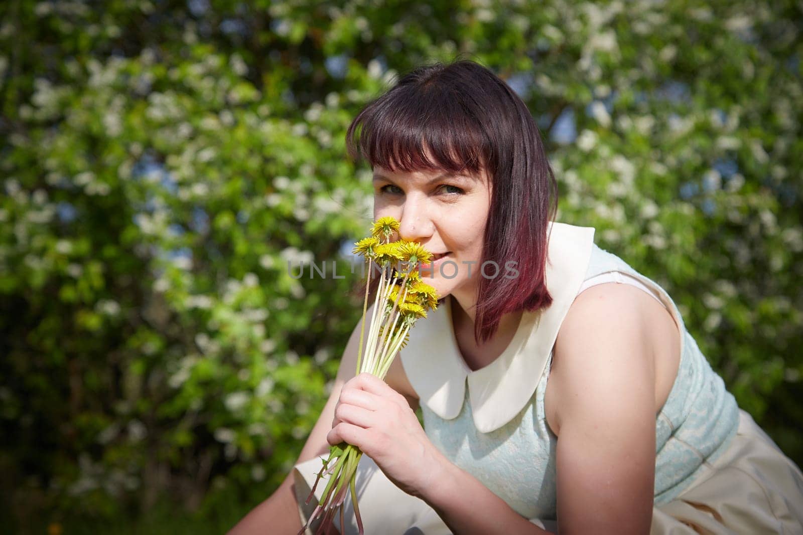 Portrait of beautiful young woman near apple trees and yellow dandelion flower in park on sunny day. Springtime