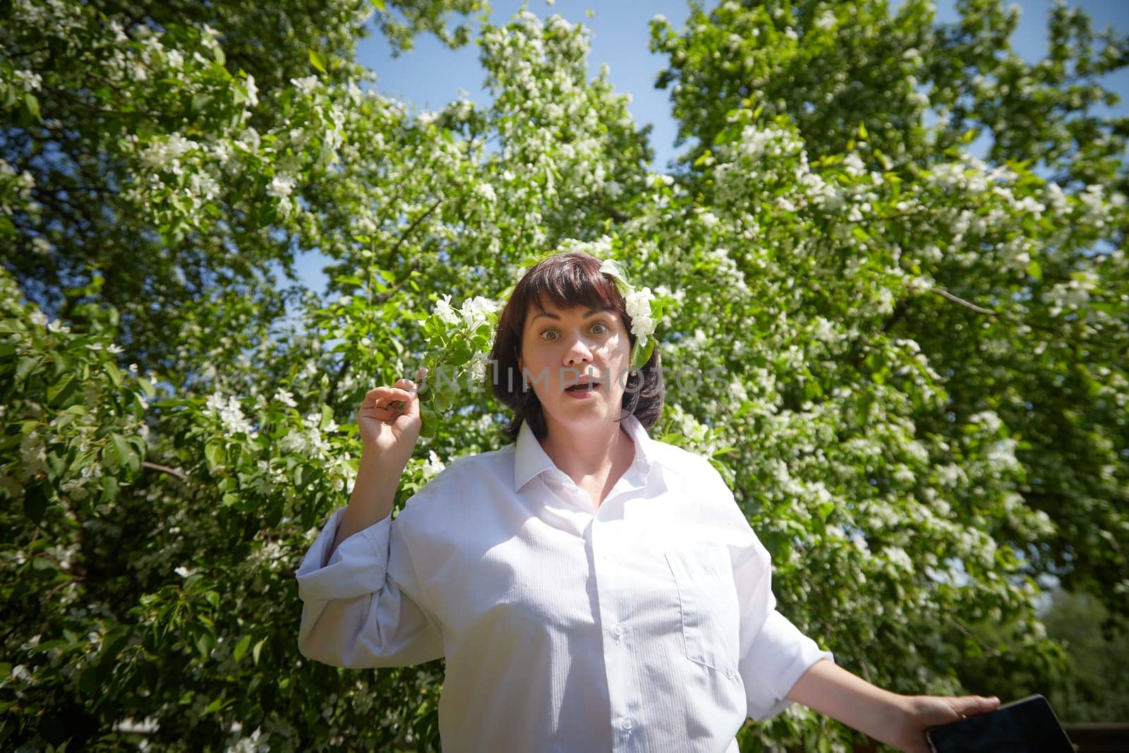 Brunette girl Using Smartphone in Blossoming Orchard in Springtime. Middle aged Woman enjoying phone among spring blossoms of apple or sakura trees