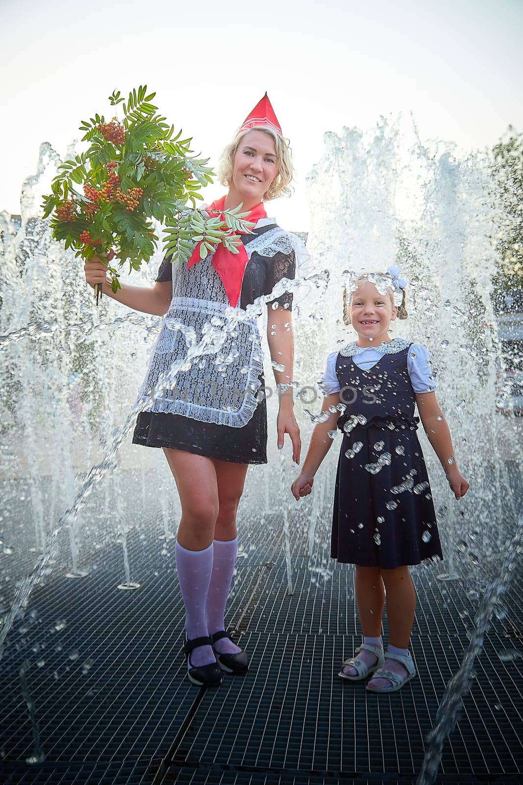Young and adult schoolgirl on September with flowers having fun near water of fontain. Generations of schoolchildren, pioneer of USSR and October girl in modern uniform of Russia. Mom and daughter