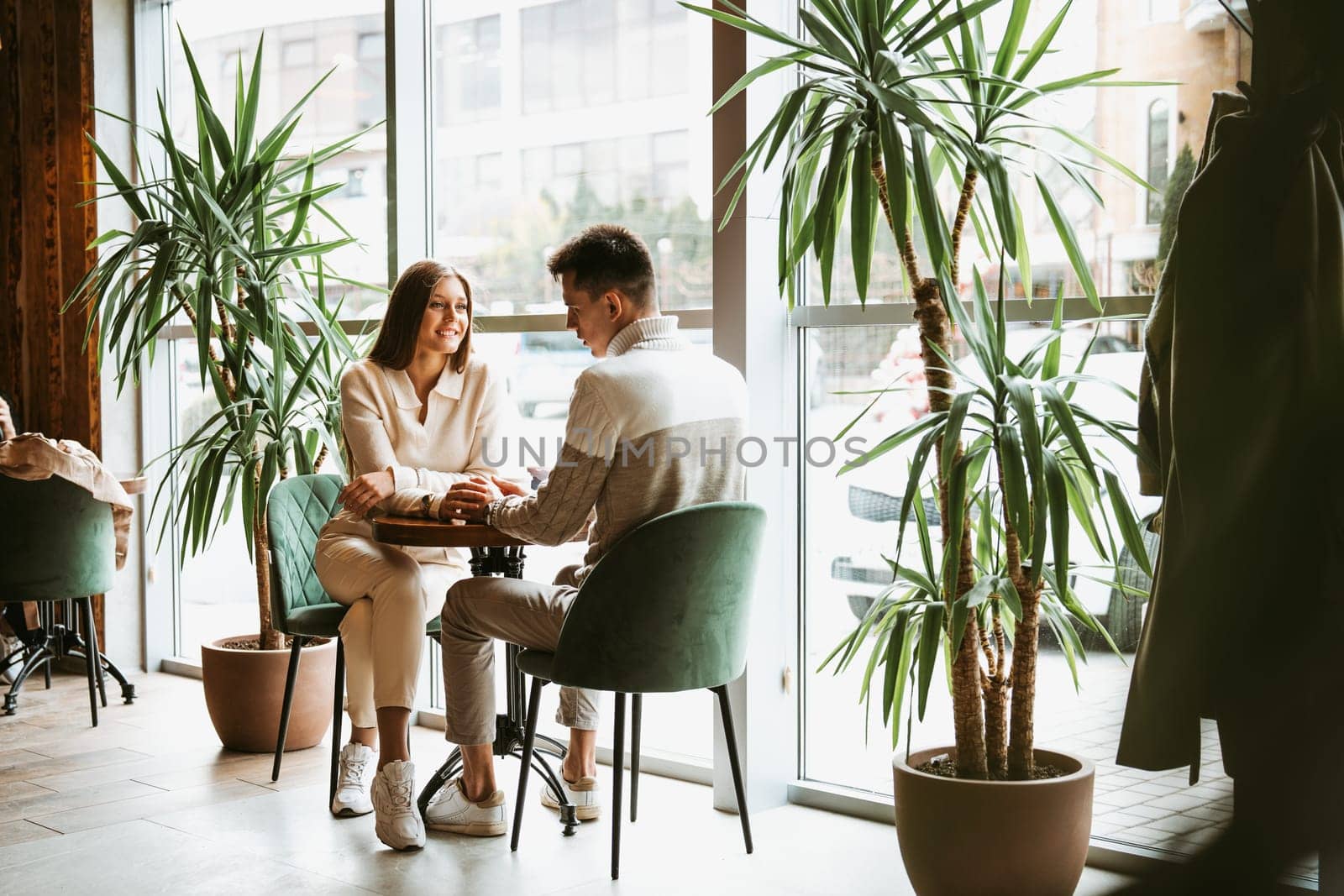 A man and a woman are seated across from each other in a sunlit cafe, holding hands and gazing into each others eyes, creating a moment of intimacy. The setting exudes warmth and tranquility, with indoor plants contributing to the serene atmosphere.