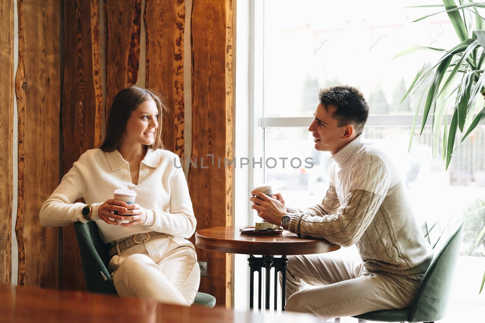 A woman and a man are sitting at a wooden table in a warmly lit cafe, their faces animated in what appears to be a relaxed conversation. The woman holds a paper cup, possibly containing coffee, as they both enjoy a comfortable moment together, surrounded by an ambiance of casual chatter.