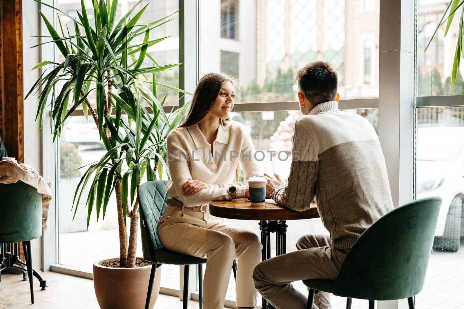 A man and woman are seated opposite each other at a small table, deeply engaged in a conversation. The man, wearing a white knit sweater, listens attentively to the woman with his hands gently clasped. The warm ambiance of the cafe, with soft natural light spilling in, adds to the intimate setting.