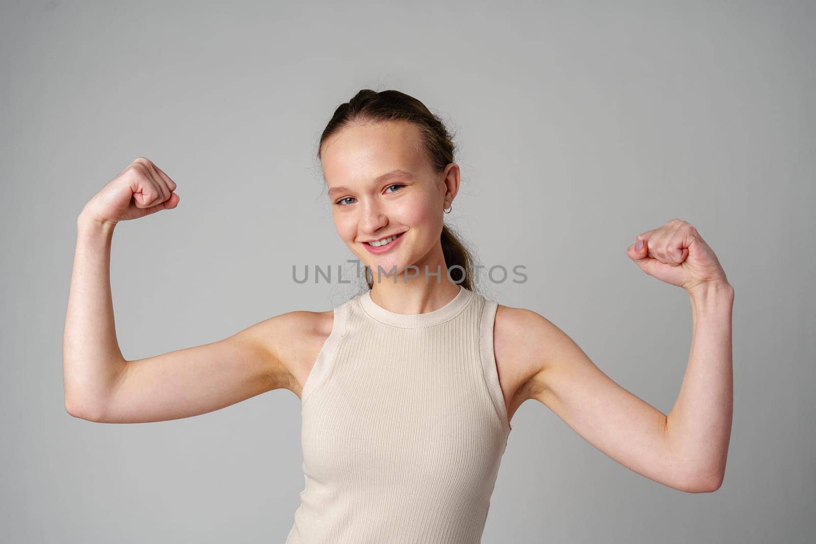 Young Woman Flexing Muscles in Beige Tank Top on gray background by Fabrikasimf