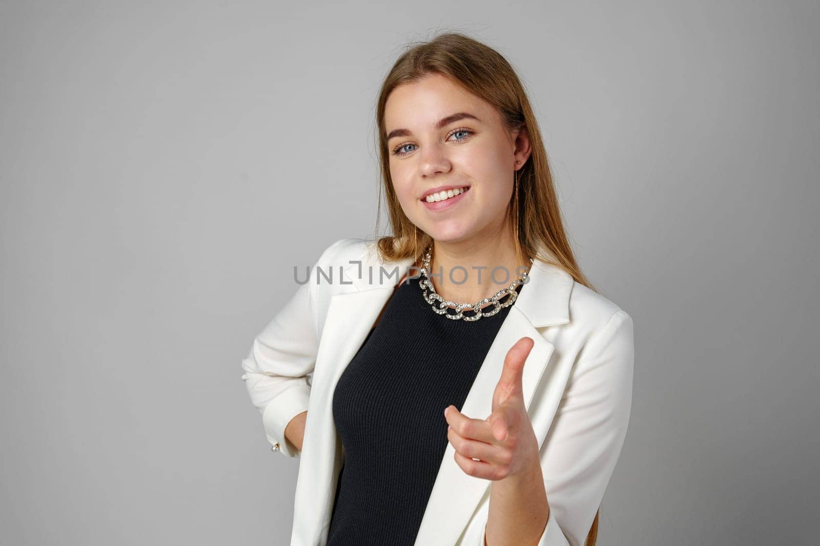 Young Woman in White Blazer Pointing at You Against a Grey Background in Studio